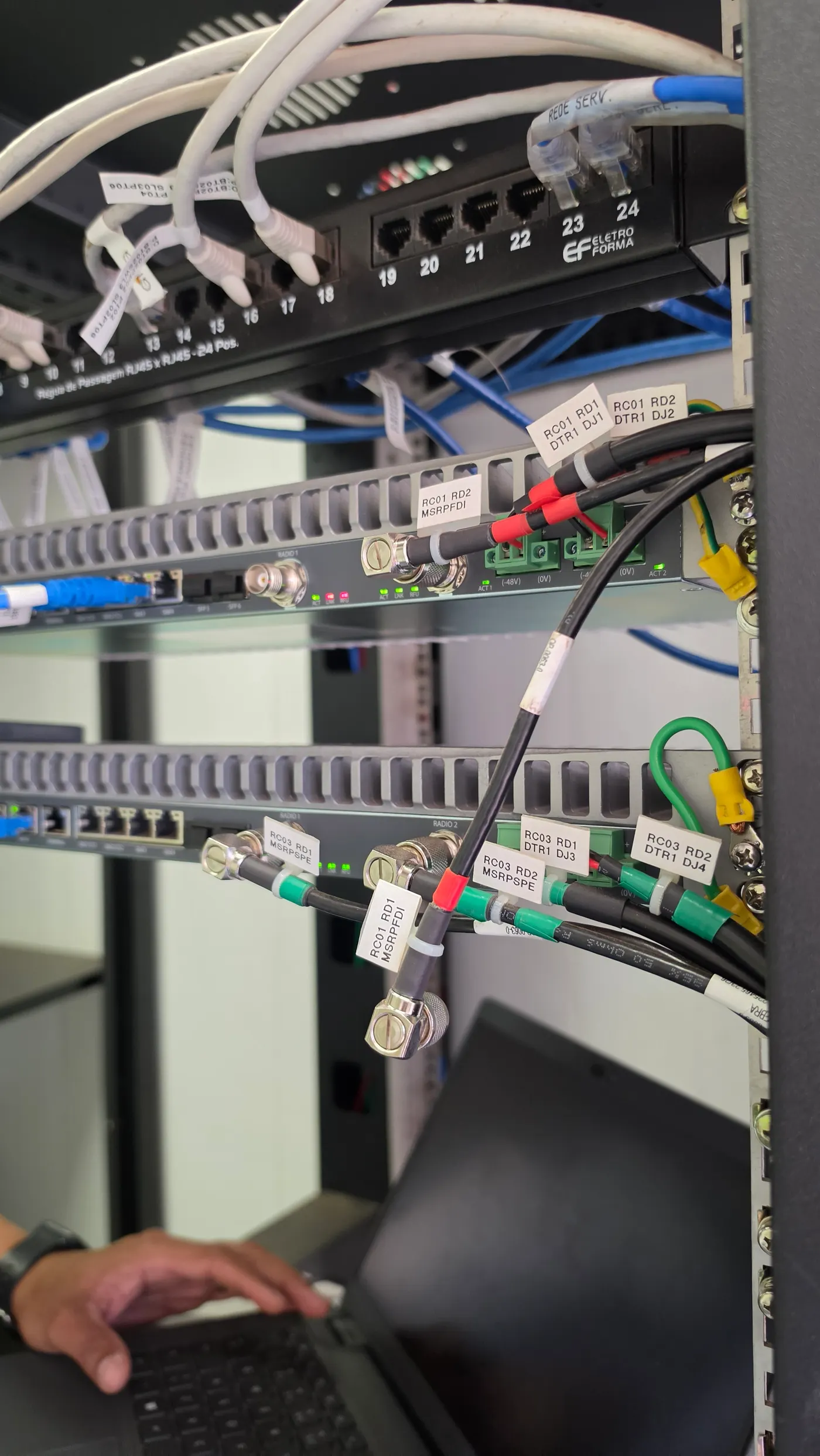 Tecnico da i3F conferindo pelo notebook o cabeamento identificado de um rack de transmissao
